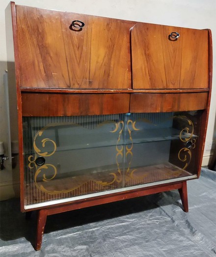 A wooden cabinet approx 110cm wide by 110 tall by 30cm deep, in a dark brown veneer. The lower part has a class shelf, with glass sliding doors that have a painted swirling design near the edges. in the middle are two short curved drawers. The top section was two drop-down doors that convert to shelves.  The base has 4 legs slightly splayed out.