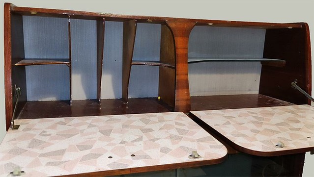 A view on the inside of the top section of the cabinet, The drop-down shelves are covered in melamine with a pink, brown and grey random large block pattern of different shapes. The rear is white but the sections of shelf inside are the same colour as the cabinet, dark brown wood. The left section is split into 4 vertical sections, with the outer section is also split horizontally, making 6 cubby holes. The right section have a horizontal glass shelf in the middle.