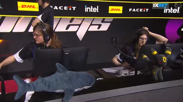 A BLÅHAJ shark plushie lies on the player desk during the ESL Impact Season 7 tournament. The blue and white stuffed shark rests on the gaming setup where the Supernova Comets team is competing, with the female pro players wearing headsets at their stations. Sponsor logos for DHL, FACEIT, and Intel are visible in the background.