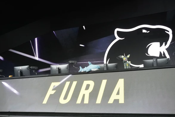 The BLÅHAJ shark sits on the main tournament stage platform at the ESL Impact event, positioned on the player setup area next to what appears to be a small mascot figure. The arena features the large 'FURIA' team logo and professional esports stage lighting, with the shark having been part of the competitive gaming environment.