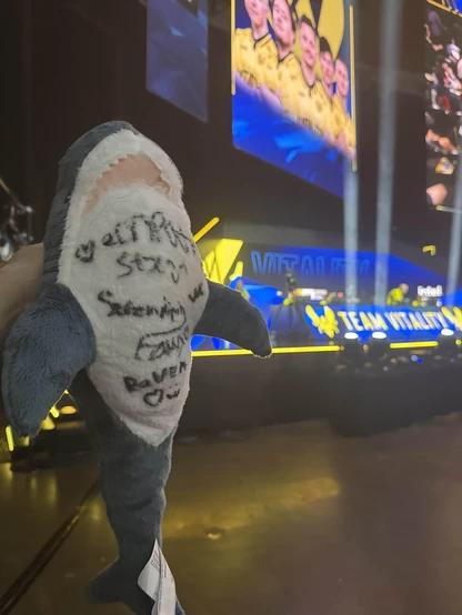 Close-up of the BLÅHAJ shark showing signatures from the Supernova Comets players written in black Sharpie on its white belly. The signatures include the team members empathy, stx, serendipity, fawx, and raven. The shark is photographed against the backdrop of the tournament venue with blue and yellow Team Vitality branding visible.