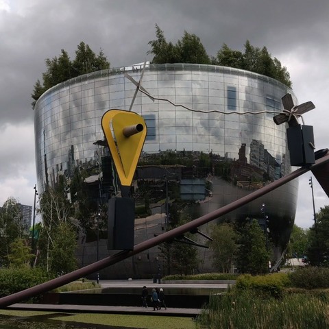 Photo of the depot of the Boijmans van Beuniningen museum, showing the vase shaped building covered with mirrors with trees on top. In front a modern sculpture with a bright yellow triangle floating in the air. | Foto van het depot van museum Boijmans van Beuningen, met het vaasvormige gebouw bedekt met spiegels en bomen erop. Op de voorgrond zweeft een moderne sculptuur met een felgele driehoek zwevend in de lucht.