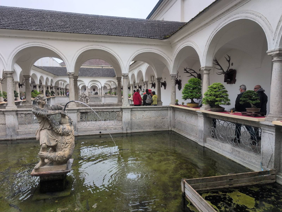 Monastary with fish ponds and a fountain in the forground. Different bonsai displayed under the arcs of the hallway. Some people in the background.