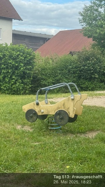 Eine Frühlingswippe für Kinder in Form eines Autos, auf grasbewachsenem Boden mit einigen Sträuchern und einem Gebäude im Hintergrund.