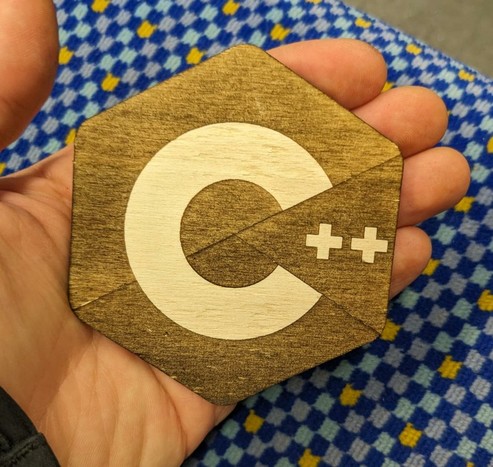 A coaster shaped in a hexagon. The C++ logo with a dark background. Held by a hand.