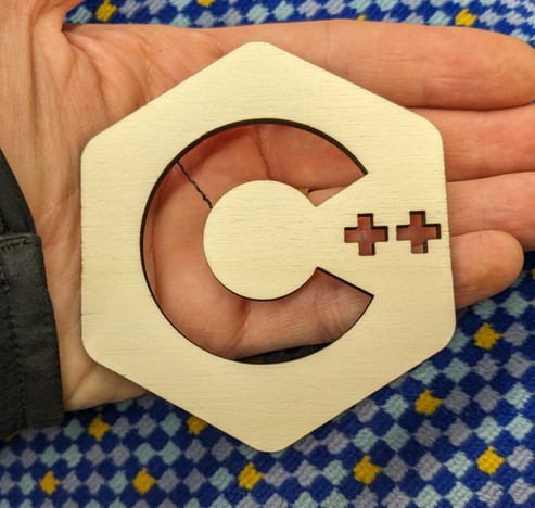 A coaster shaped in a hexagon. The C++ logo is cutted out. Held by a hand.