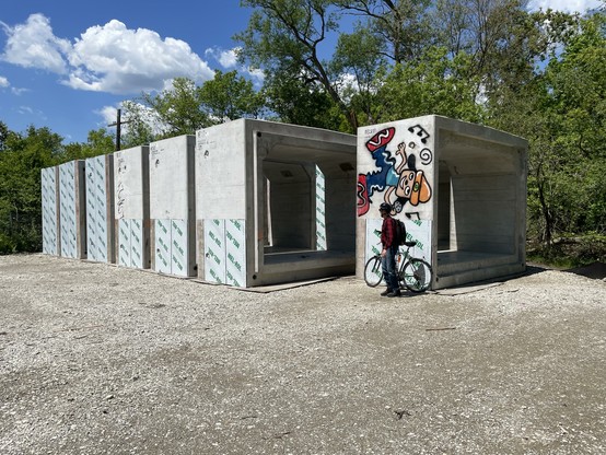 A row of seven rectangular concrete tunnel section sit in a gravelled area in the woods, waiting to be installed under a nearby train track.  In front of them stands a man with a bicycle, about half their height.