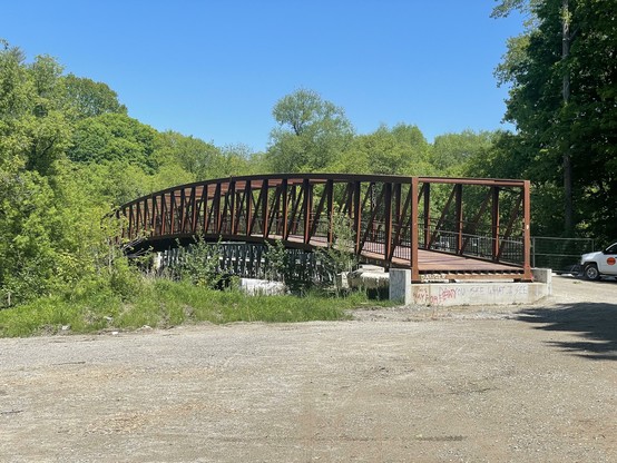 A rust-coloured gently-arched bridge spans a narrow river in a green park.  The bridge is missing one end.  Barely visible behind it is a grey flat temporary bridge.