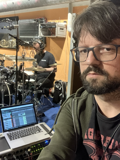 Selbstporträt vor einem Laptop mit Recording-Software, im Hintergrund ein Schlagzeuger beim Spielen