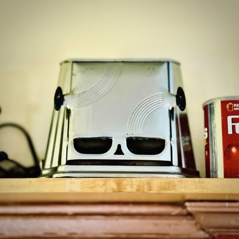 A small, silver kitchen appliance sits up on a high wooden shelf next to a Folger's coffee can, the appliance's boxy metal frame pierced with holes resembling two large eyes, a small nose, and a thin line for a mouth, along with two black handles along the sides resembling ears.