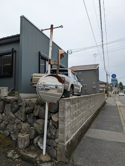 The seaside can be seen reflected on a road mirror opposite to the water. Behind the roadmirror there is a wall supporting an elevated house and a pickup truck.