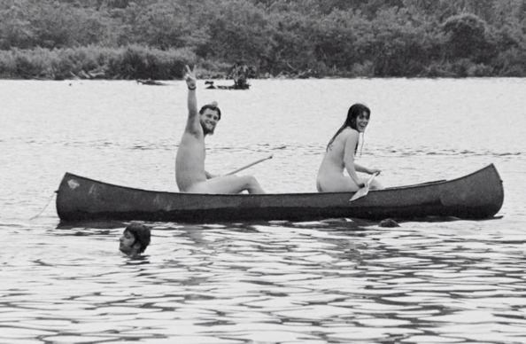 I can see this is a black and white photograph showing three people enjoying time at a lake or pond. Two people are sitting in a canoe on the water - one person is raising their hand in a cheerful gesture while the other is holding a paddle. A third person is swimming in the water nearby. The scene is set against a backdrop of trees and appears to show people having a relaxed, joyful time outdoors. [by claude ai]