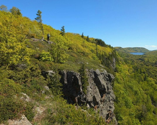 Jeff and Tonttu are small figures on a high slope coming down form left to right, covered in bright yellow-green leaves of stunted trees on the exposed face of mountaintop. The slope meets a steep cliff of dark stone that drops away into the valley below in dramatic fractured vertical shapes. A few isolated evergreens stick up through the low deciduous trees on the slope, and full-sized mature forest stands in the valley below. Lake of the Clouds can still be seen as a distant splash of blue far back in the valley.