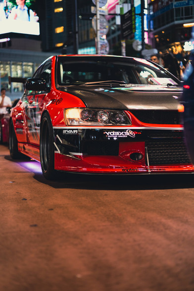photo of a Mitsubishi Lancer Evolution VII tuning car from the front, with its headlights in focus. It's night on y lively and well illuminated street.