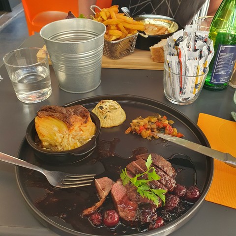 A plate with duck breast and multiple sides (gratin potatoes, souffle, vegetables) in the foreground, another with a backed camembert further in the back.