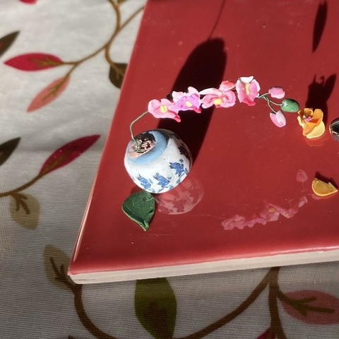 A tiny ceramic vase with blue floral patterns holds a delicate arrangement of pink orchid flowers in a single spray. The vase sits on a glossy red tile, surrounded by scattered yellow petals, and the reflection of the model can be seen in the gloss. It has a patterned fabric tablecloth in the background.