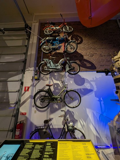 Five motorized bikes hang on the wall at the Malmo House of Technology. They come in multiple colors. The bottom (earlier ones) clearly reflect their origins as bicycles with small added internal combustion engines. In the bottom of the photo, the interpretive panel is visible but illegible.