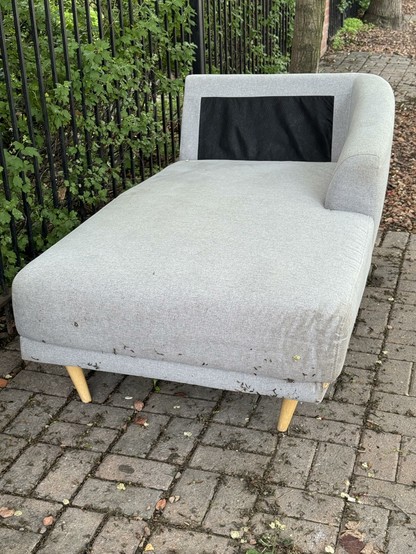 A cream chaise lounge chair is sitting on the sidewalk outside. You can see a wrought iron fence and some leafy green bushes behind the fence to the left of this shot. The futon is sitting on a portion of sidewalk made of brick-shaped stones that are partially covered in moss and dead leaves.