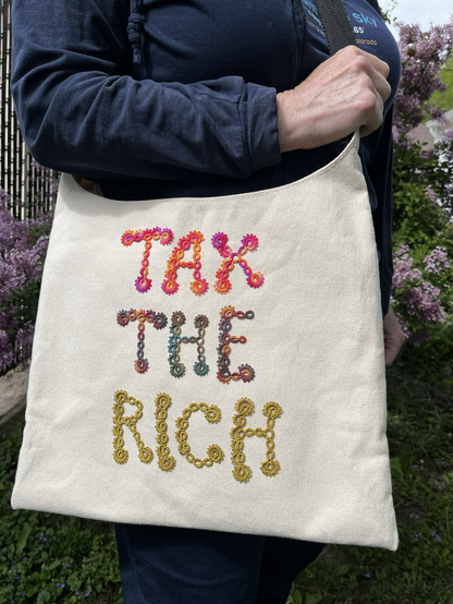 Off white canvas bag with colorful tatted lace letters on it spelling out “Tax the Rich”.