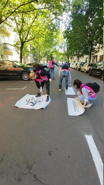 Ein Foto von einer Straße, auf der 4 Menschen in pinken Warnwesten tagsüber einen Radweg selbermachen. Mit Schablonen sprühen sie Fahrradwegmarkierungen und -symbole auf die Straße.
