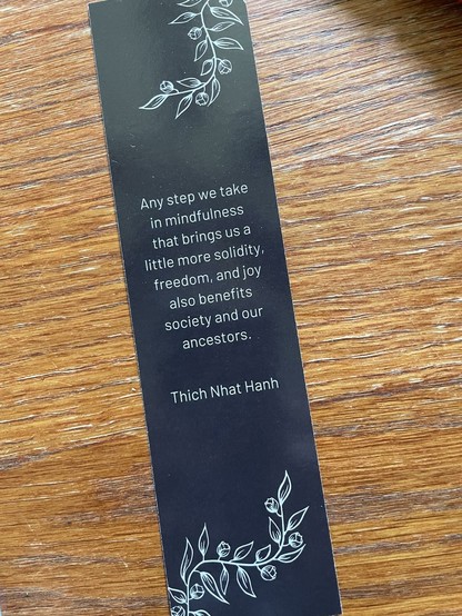 A black bookmark on a wooden table. In white outline is flowering vine on top and bottom edges. In center is Thich Nhat Hanh quote:

Any step we take 
in mindfulness
that brings us a 
little more solidity,
freedom, and joy
also benefits
society and our
ancestors.