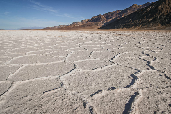 A vast salt flat with polygonal, cracked patterns stretches across the foreground under a clear blue sky. The textured white surface contrasts with the rugged, sunlit mountains rising on the right side of the image, creating a stark and dramatic desert landscape.