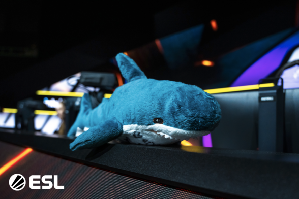A plush blue shark toy (Blåhaj) rests on the front edge of an esports stage at the ESL Impact Season 7 Grand Finals. Behind it, colorful stage lights and gaming setups are visible, with the ESL logo in the lower-left corner.