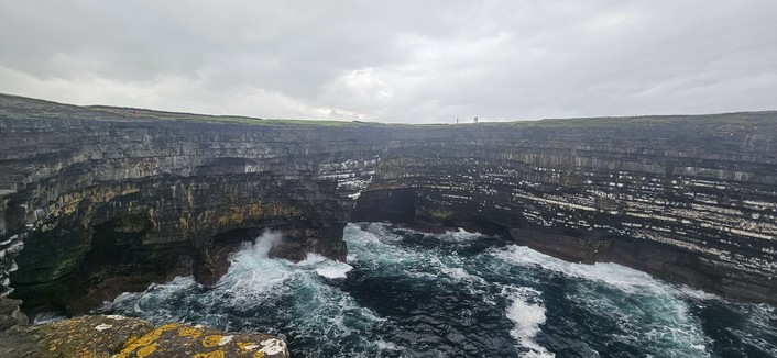 A dramatic view of steep, layered sea cliffs with dark rock faces, as waves crash against the base under a cloudy sky. The landscape is wild and rugged, with the sea appearing turbulent below