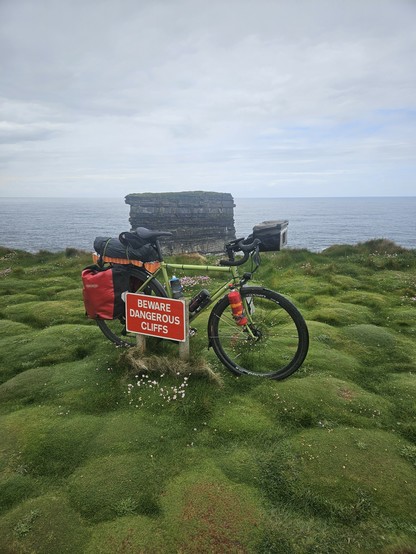 A touring bicycle loaded with panniers and gear is parked on lush, uneven green grass near the edge of a cliff. A red sign attached to the bike reads “BEWARE DANGEROUS CLIFFS.” In the background, a large isolated sea stack rises from the ocean