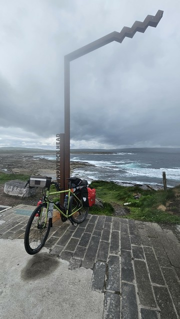 A touring bicycle is parked beneath a modern, zigzag-shaped metal sculpture labeled “DOWNPATRICK HEAD.” The bike is loaded with panniers and gear, and the dramatic coastline with waves breaking on rocks is visible in the background under a cloudy sky4