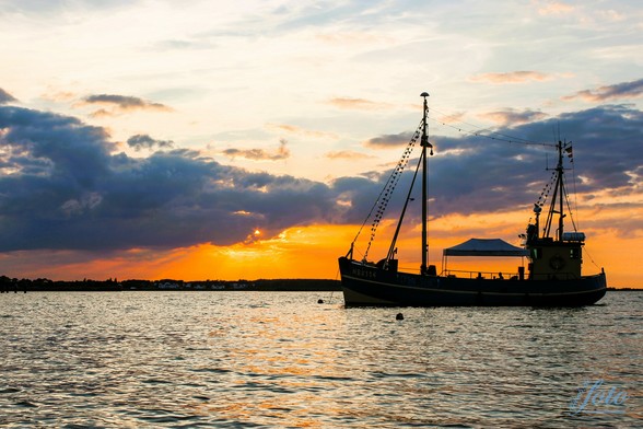 Ein Sonnenuntergangsfoto zeigt im Vordergrund Wasser mit kleinen Wellen, im rechten mittleren Bereich ein Boot im Sonnenuntergang. Dahinter ein paar Dramawolken, welche von der untergehenden goldgelben Sonne angestrahlt werden.