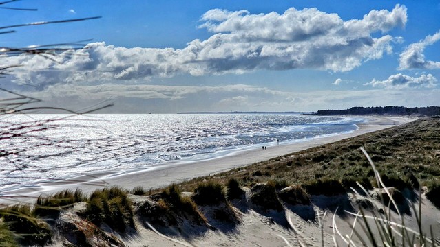 A wide view of an expansive beach on the East Coast of Ireland, featuring bright, sparkling water, a wide sandy shore, and grassy dunes in the foreground. Two small figures are walking along the water's edge, with a distant landmass visible. The sky is partly cloudy, and the sunlight creates reflections on the sea.