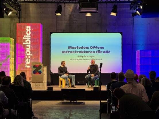 A stage at re:publica - two men sitting in front of a white slide with the words "Mastodon: Offene Infrastrukturen fur alle".