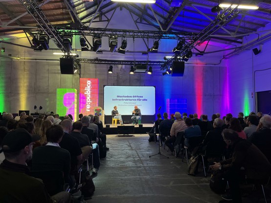 A long shot of a stage at re:publica - in the distance, two men sitting in front of a white slide with the words "Mastodon: Offene Infrastrukturen fur alle". The photo is taken along the central aisle, with rows of attendees on either side.
