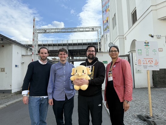 Members of the Mastodon team, 3 men and a woman, holding a beige plush Mastodon mascot, near some industrial looking buildings. The sky is blue with white fluffy clouds.