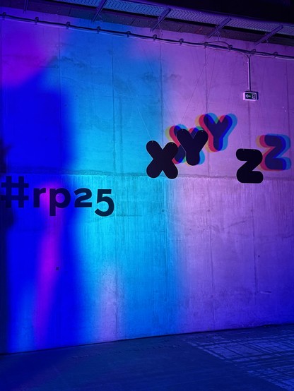 The re:publica hashtag rp25 on a concrete wall lit with neon blue and pink lights, with the letters X, Y, and Z hanging and casting shadows nearby.