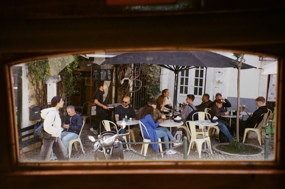 Vue depuis la fenêtre rectangulaire d’un tram : des personnes sont assises en terrasse sous un jour de soleil, profitant d’une boisson