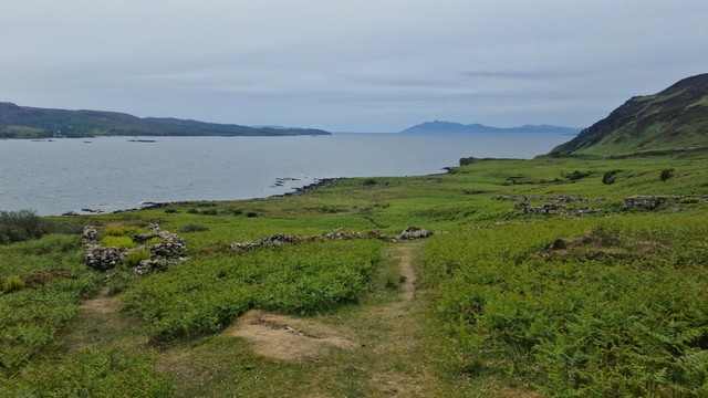 Crumbling stone ruins lie amid thick vegetation on a sloped hillside leading down to the lochside. Numerous sheep are dotted around. The sky is cloudy above. The trail leads down through the middle of the scene.