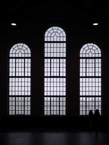 The silhouettes of two women stand out in front of a decorated window which is part of a group of three. The windows all have small glass panels and a circular top. The rest of the scene is pitch black.