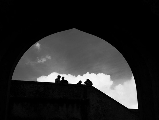 The silhouettes of four people are visible on top of a structure and stand out in front of a white cloud. The top and bottom of the image are pitch black, as the arch structure blocks all the light.