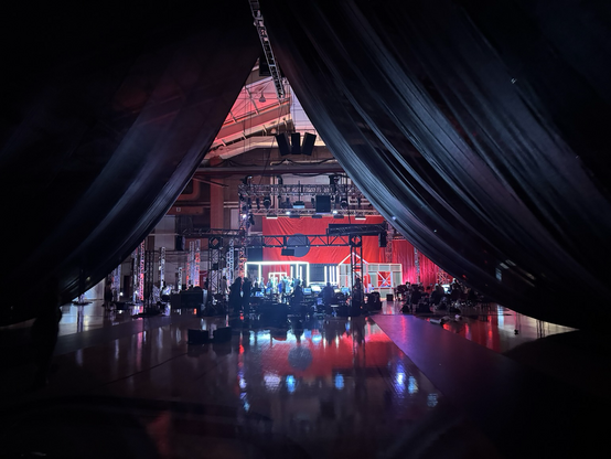 A photo of the stage for "Sensorium Ex" as shot from way back in the house. There are dark black drapes over the centre of the room, followed by bright lights up ahead.  The floor is reflective. The stage has a red background colour.