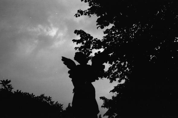 Underexposed and quite dark black and white film photo of a statue of an angel against a cloudy sky and surrounded by greenery