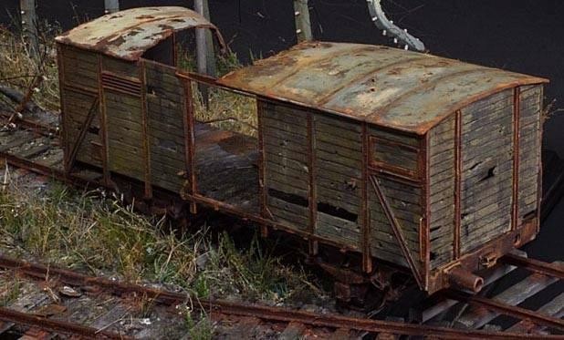 This image captures a derelict freight wagon resting on rusted, overgrown railway tracks, evoking a profound sense of abandonment and decay. The wagon’s structure is a haunting testament to its age. Its wooden panels, warped and splintered, bear countless scars—gaping holes and deep cracks that speak of prolonged exposure to the elements. The metal framework is cloaked in layers of rust, an oxidized palette of ochre and deep brown that amplifies its weathered appearance. The roof, with its uneven corrugation, is dotted with patches of corrosion, giving it a precarious, fragile look.
