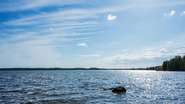 A vast expanse of water under a bright blue sky, with scattered clouds. The sunlight creates glittering reflections on the water's surface. A dark rock protrudes from the water in the foreground, and tree-lined shores are visible in the distance on the right side of the image.