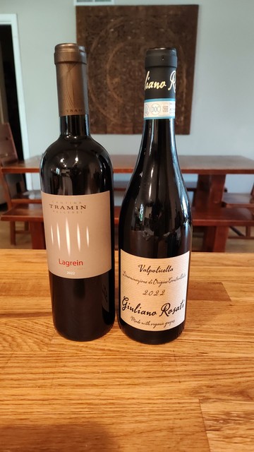 Two bottles of wine rest on a wooden counter with a table and wooden wall hanging behind. They are both Italian, one is a pure Lagrein and the other is a Valpolicella.