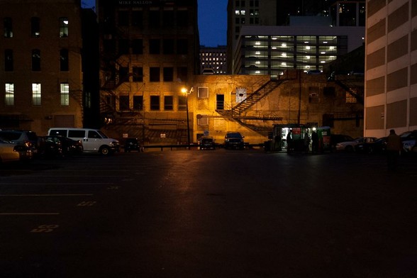 A nearly empty parking lot with buildings surrounding it at night. A single sodium lamp illuminates the back of the parking lot casting shadows and light on the nearby buildings. Further behind other buildings with lights in their windows are seen (save one, to the left of the sodium lamp)