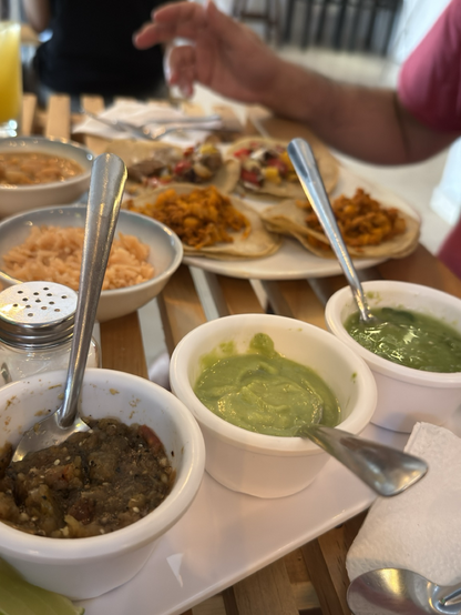 Fotografía de unos recipientes blancos con salsa roja, guacamole y salsa verde. En la mesa hay un salero y unos platos hondos con arroz y frijoles charros y un plato con cinco tacos.