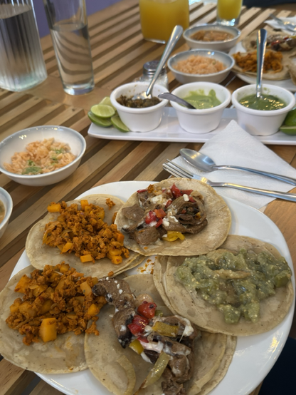 Fotografía de un plato blanco con 5 tacos, dos de chorizo de soya, dos de asada ande seitán con pimientos moreno amarillos, rojo y queso vegetal, un taco de chicharrón en salsa verde, en la mesa de madera también hay unos platos de arroz y frijoles charros, unos recipientes blancos con salsa roja, salsa verde, guacamole y limones. También hay unos cubiertos y una servilleta al lado del plato grande.