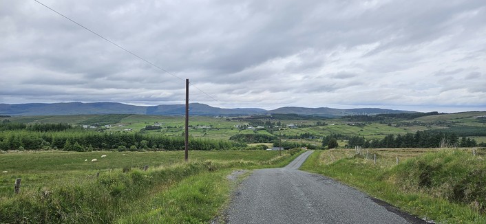 A narrow rural road winds downhill through open countryside, flanked by grassy fields and scattered sheep. In the distance, clusters of houses and green hills stretch beneath a cloudy sky.