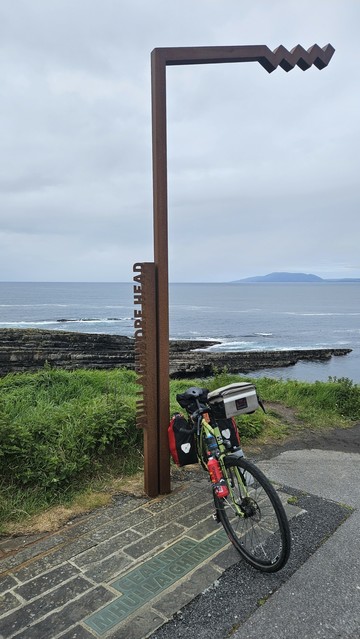 A touring bicycle loaded with bags is parked beside a modern, rust-colored metal sculpture marking "Mullaghmore Head." The sculpture stands on a stone path overlooking dramatic rocky coastline and the Atlantic Ocean, with a distant island visible under a cloudy sky.